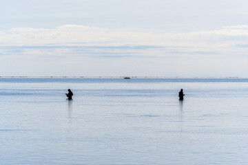 Silhouettes of two fishermen fishing standing waist-deep in the calm Mediterranean Sea at the mouth of the Ebro River, Trabucador beach on the horizon and a boat with three fishermen fishing.