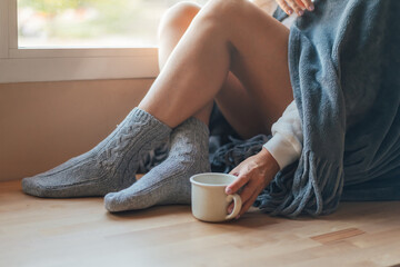 Winter Moments.Young Woman Relaxing By The Window, Sipping A Warm Cup Of Coffee Or Tea,Wearing Wool Socks And A Cozy Blanket, Capturing The Essence Of Comfort And Nostalgia On A Cold Afternoon At Home