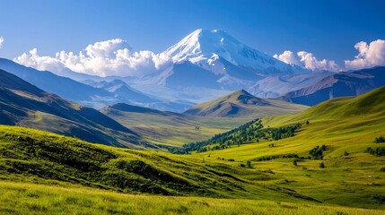 Fototapeta premium Majestic snow-capped mountain peak rising above rolling green hills and valleys, with fluffy clouds in a clear blue sky.