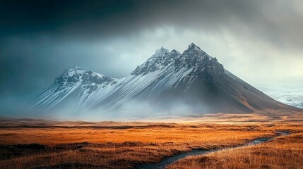Obraz premium Majestic snow-capped mountain peaks rise above a field of dried grass and a small stream in Iceland with a dramatic sky.
