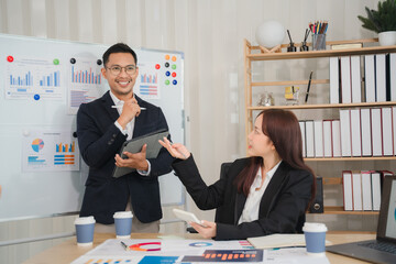 Business Meeting: A confident businessman presents a digital tablet to a female colleague during a professional meeting, with charts and graphs on a whiteboard behind them.