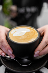 A close-up of hands holding a black cup with a creamy latte, featuring intricate foam art. background is blurred, highlighting the focus on the coffee. Perfect for themes of warmth and coffee culture.