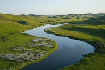 Obraz premium Aerial view of a calm, serene river snaking through a lush green valley, reflecting the clear sky above, bordered by rolling hills and patches of wildflowers blooming along the water’s edge