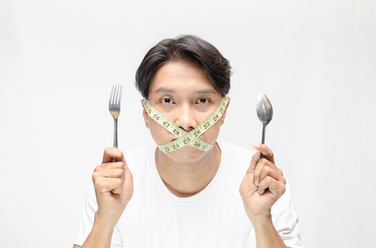 a diet concept illustrated by young asian man with unhappy expression covering his mouth with a measuring tape. eating disorder concept. extreme diet concept. man holding spoon and fork hungrily.