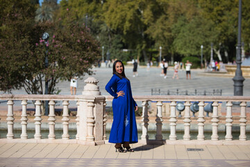 Fototapeta premium young and beautiful moroccan woman in blue abaya, typical arabian costume, walking along the canal in the square of spain in Seville, a world heritage site, on a sunny day. Travel concept.