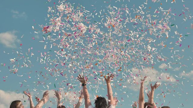 Friends celebrating joyfully outdoors, throwing colorful confetti under a blue sky. Arms raised in excitement, surrounded by a cheerful atmosphere