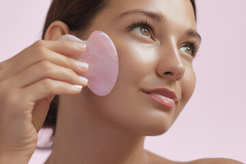 A young woman gently massages her face with a rose quartz gua sha tool, showcasing her glowing skin and relaxed expression. Perfect for beauty, skincare, and wellness promotions focusing on natural