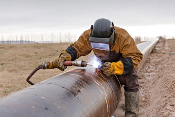 A welder meticulously joins sections of a large pipeline, sparks flying, as the sun sets over the rugged terrain, highlighting the labor involved in pipeline construction.