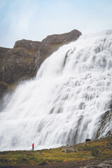 A hiker in a red jacket dwarfed by the massive Dynjandi waterfall in Iceland's Westfjords region.