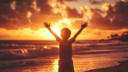 Fototapeta premium A boy on a beach with his arms raised high, silhouetted by the setting sun, as the ocean waves gently roll in and the sky bursts with vibrant sunset colors.