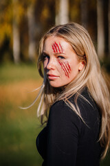 A woman outdoors with fake bloody claw marks on her face, possibly in costume for Halloween or a...