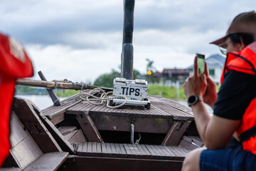 "TIPS" box on boat for river cruise, with tourist in life jacket taking photo on cell phone. Illustrating that Tip Boxes are everywhere.