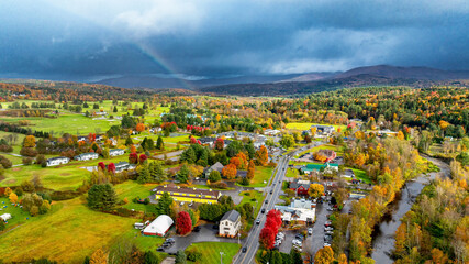 Scenic autumn landscape of a quaint USA village. A picturesque village in the USA showcases vibrant autumn colors under a dramatic sky, with a hint of rainbow amidst rolling hills.