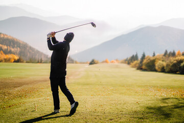 A golfer in mid-swing on a scenic golf course with mountains and autumn trees in the background, under soft sunlight..