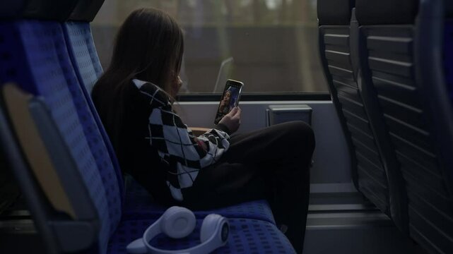 teenager is sitting on a blue patterned seat inside a train and listing to the music with headphones