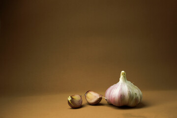The image shows garlic on a brown background. On the right is a whole head of garlic, and on the left are two individual cloves. The head of garlic is a light purple color, and the cloves are a darker
