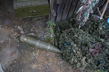A scene of military equipment and camouflage netting in a neglected area. An artillery shell and a green box with Cyrillic text are visible. Weeds grow among the debris, showing nature reclaiming the 