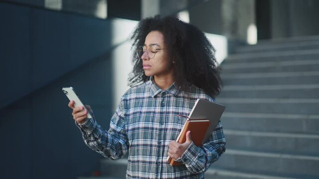 Confused young african american woman student looking up at university signs and map trying to find his way using mobile phone at urban buildings alone Lost in the campus Digital navigation