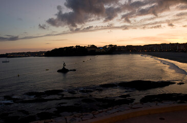 The sun is setting on the horizon of a city beach, with a couple standing on rocks in the foreground