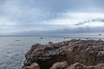 Small fishing boats floating on a cloudy day at sea near rocky coastline