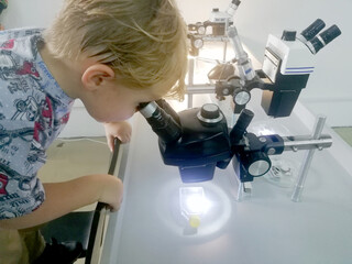 Interesting sample. Creative happy merry boy using microscope while smiling and wearing lab coat....
