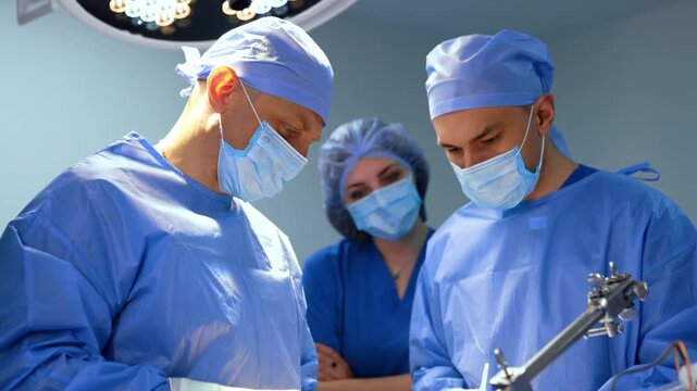Male Caucasian doctors look attentively at the patient on operating table. Surgeons consult at neurosurgery. Female nurse stands at backdrop.