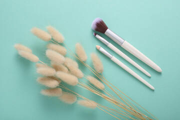 Makeup brushes with lagurus flower on mint green background. Top view. Beauty concept