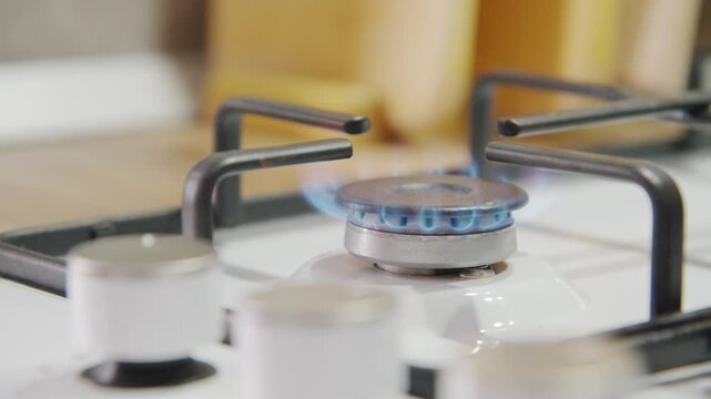 A gas stove burns with a blue flame, and an out-of-focus hand in the foreground turns the knob. Gasification of housing