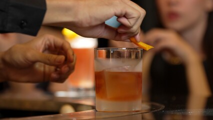 Bartender squeezing orange peel smelling over glass cocktail whiskey putting into Old fashioned on top of rock ice making special drink advertisement alcohol on Paloma and guest background. Vinosity.