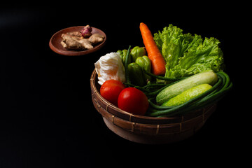 arrangement of vegetables commonly used in indonesian dishes, including tomatoes, carrot, cucumbers, long beans, pumpkin, cabbage, and lettuce. Put on traditional bamboo basket, solid black background