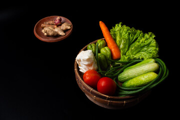 arrangement of vegetables commonly used in indonesian dishes, including tomatoes, carrot, cucumbers, long beans, pumpkin, cabbage, and lettuce. Put on traditional bamboo basket, solid black background