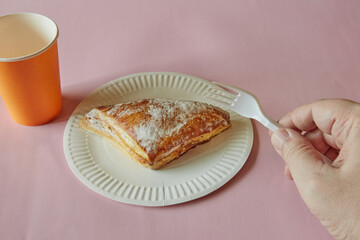 A hand holding a disposable fork over a triangle of puff pastry with sugar sprinkle on a disposable plate on a pink background, fast food, street food.