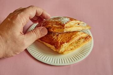 A hand picking up a triangle of puff pastry with sugar sprinkle from a disposable plate on a pink background, fast food, unhealthy eating