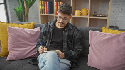 A contemplative young hispanic man with glasses and a moustache writing in a notebook while sitting on a gray couch amid colorful pillows.