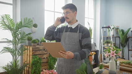 Handsome hispanic man with a moustache using a phone and holding a clipboard in a flower shop interior.
