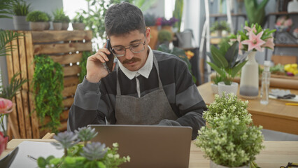 Hispanic man multitasking in a flower shop, using a laptop and phone amidst vibrant floral arrangements.