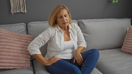 Fototapeta premium Elderly woman with short blonde hair in a white shirt experiencing pain while sitting on a grey sofa in a cozy indoor living room setting.