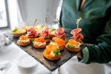 selection of gourmet canapés on a slate serving platter, featuring smoked salmon, cured meats, and garnishes, held by a person in a green sweater. Perfect for upscale catering and food presentations.