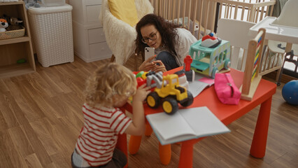 Woman photographing toddler playing with toys on red table in cozy bedroom surrounded by playthings and crib, capturing a joyful family moment in a warm home setting.