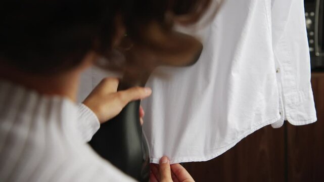 Woman steaming a white shirt at home with a handheld steamer. Clothing care, wrinkle removal, household chores, clothing care