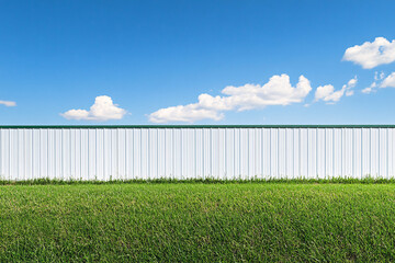 Fototapeta premium white metal fence standing behind a vibrant green lawn with a clear blue sky and few clouds creating a peaceful outdoor setting 