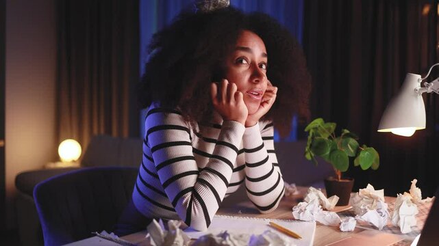 Curly african american woman student thinks questioningly about difficult choice choose imagine plan in mind has doubts create idea at desk between crumpled papers with failed ideas at late night