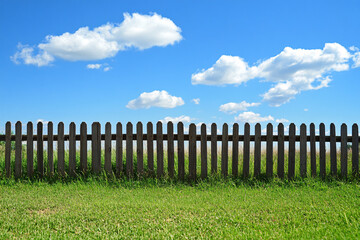 Obraz premium A simple wooden fence stretches across a green field under a bright blue sky with scattered clouds, representing a calm and sunny rural setting 