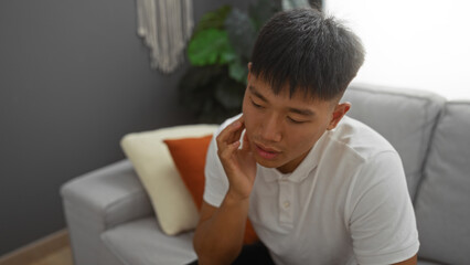 Handsome young chinese man sitting indoors in a modern living room wearing a white shirt looking contemplative with his hand on his chin