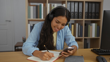 Young woman studying in a library with headphones, smiling while writing in a notebook and using a phone, surrounded by bookshelves and files