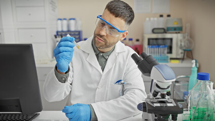 A young hispanic man analyzes a sample in a laboratory setting, equipped with safety glasses and a lab coat.