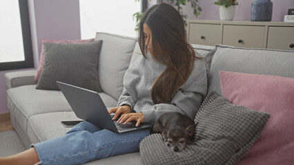 A young woman works on a laptop with her chihuahua resting beside her on a couch in a cozy living room.