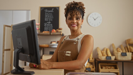 Woman working in a bakery with curly hair and wearing overalls standing behind counter and smiling...