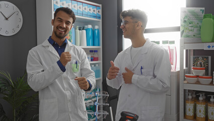 Two male hispanic pharmacists in lab coats smiling and talking together in a well-stocked pharmacy interior, showcasing a friendly and professional work environment surrounded by various products.