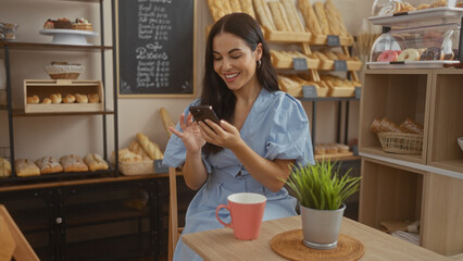 Attractive hispanic brunette woman smiling while using smartphone in a bakery shop with various breads and pastries, seated at a table with a coffee mug and plant, creating a cozy indoor ambiance.